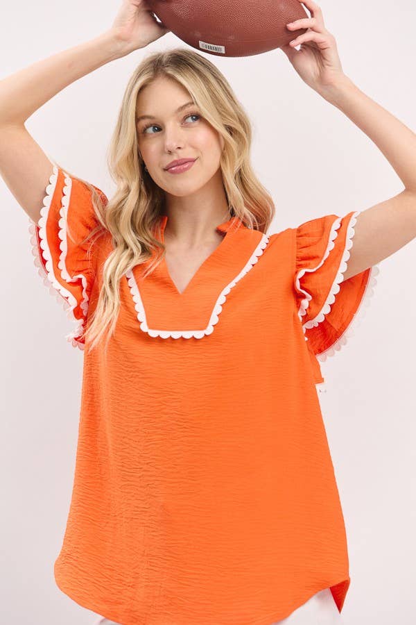 Woman wearing an orange blouse with white trim, holding a football above her head against a plain background.