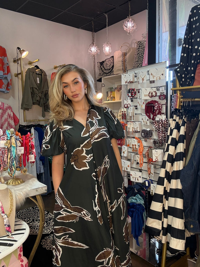 A woman modeling a dark green short-sleeve maxi dress with a brown and white abstract floral pattern, standing inside a boutique shop surrounded by clothing racks and display walls.