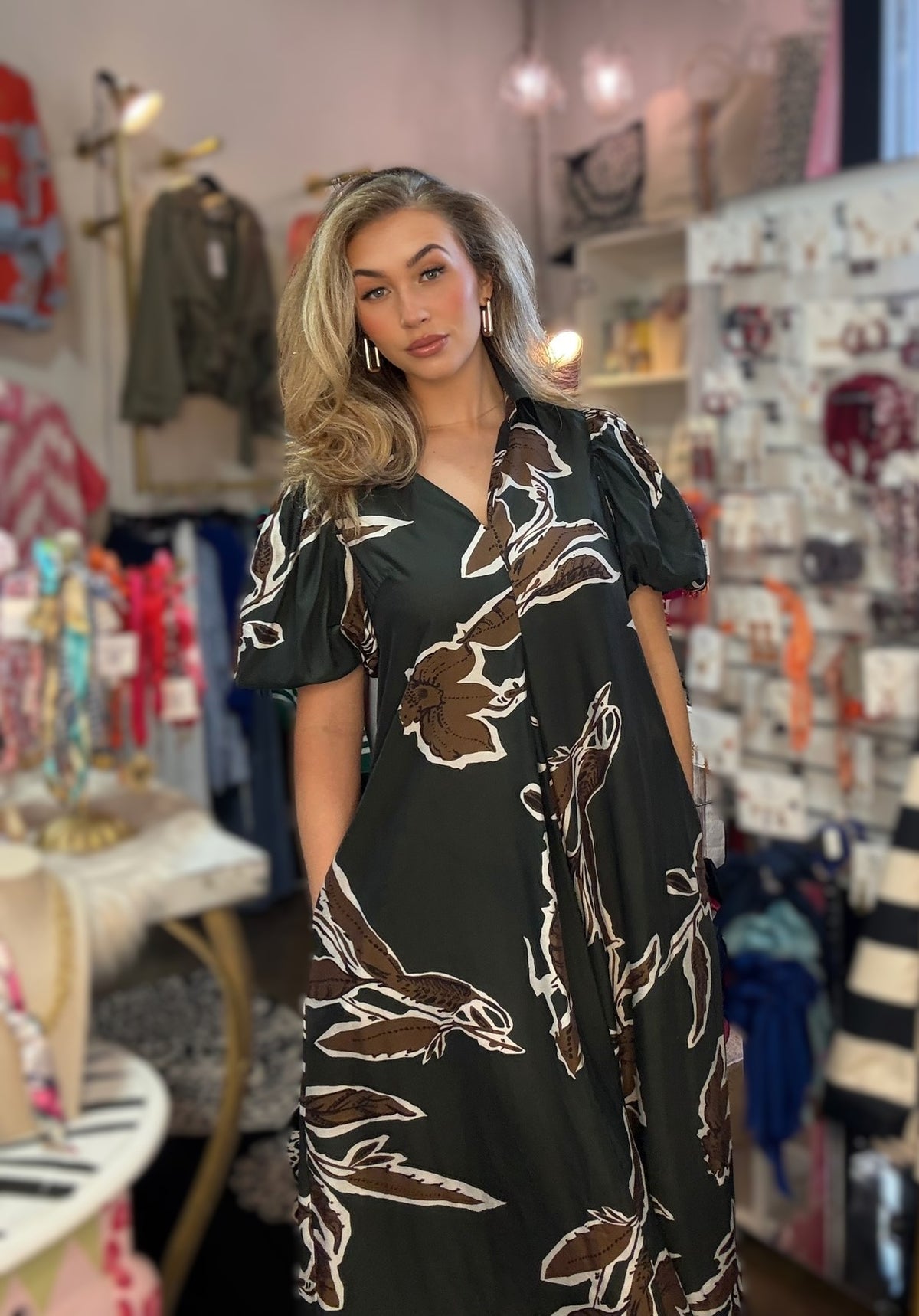 A woman modeling a black maxi dress with brown and white floral patterns in a boutique setting.