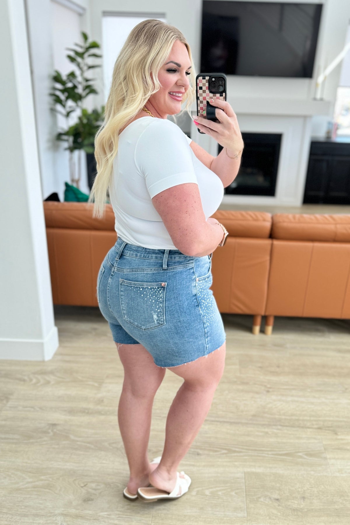 Woman taking a mirror selfie wearing a white t-shirt and denim shorts in a living room.