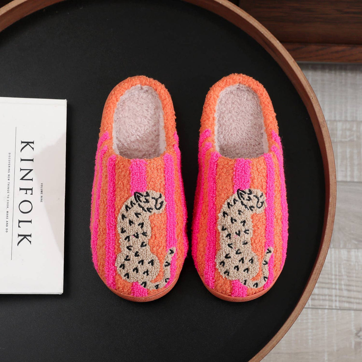 A pair of fuzzy slip-on house slippers with bright pink and orange vertical stripes and a leopard print motif on the front, displayed on a black tray next to a white book.