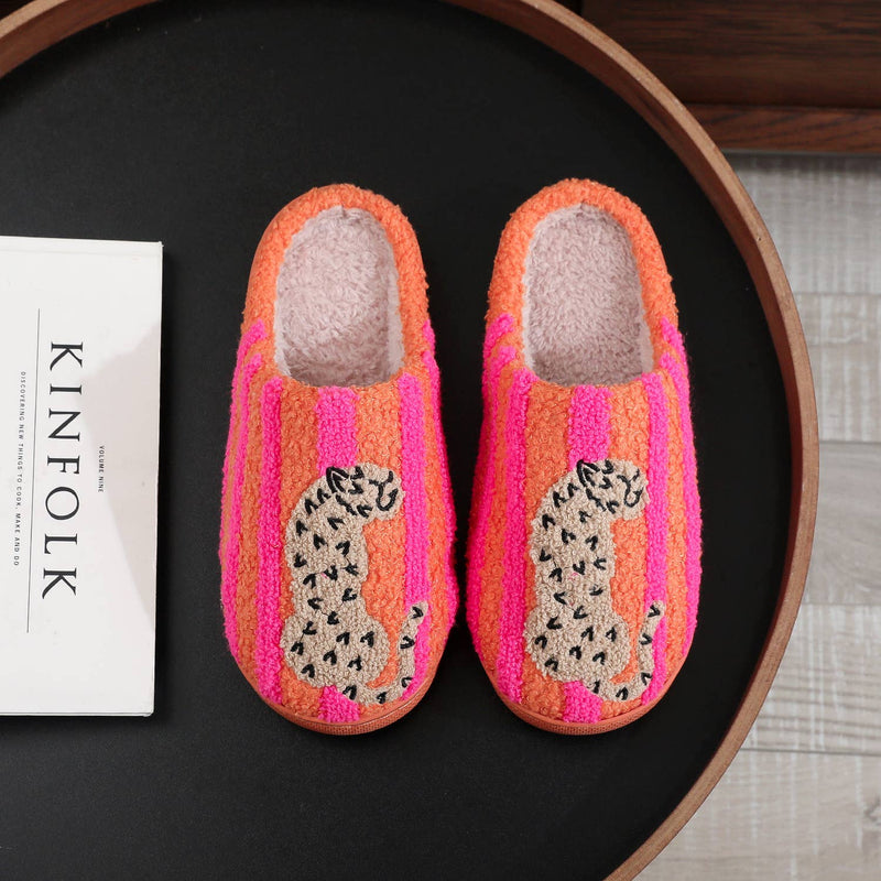 A pair of fuzzy slip-on house slippers with bright pink and orange vertical stripes and a leopard print motif on the front, displayed on a black tray next to a white book.