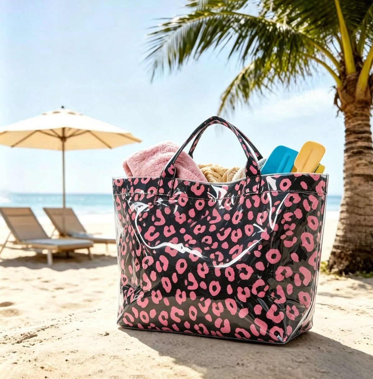 A large glossy tote bag with a pink leopard print pattern on a black background, filled with towels and beach accessories, sitting on a sandy beach with lounge chairs and a palm tree in the background.