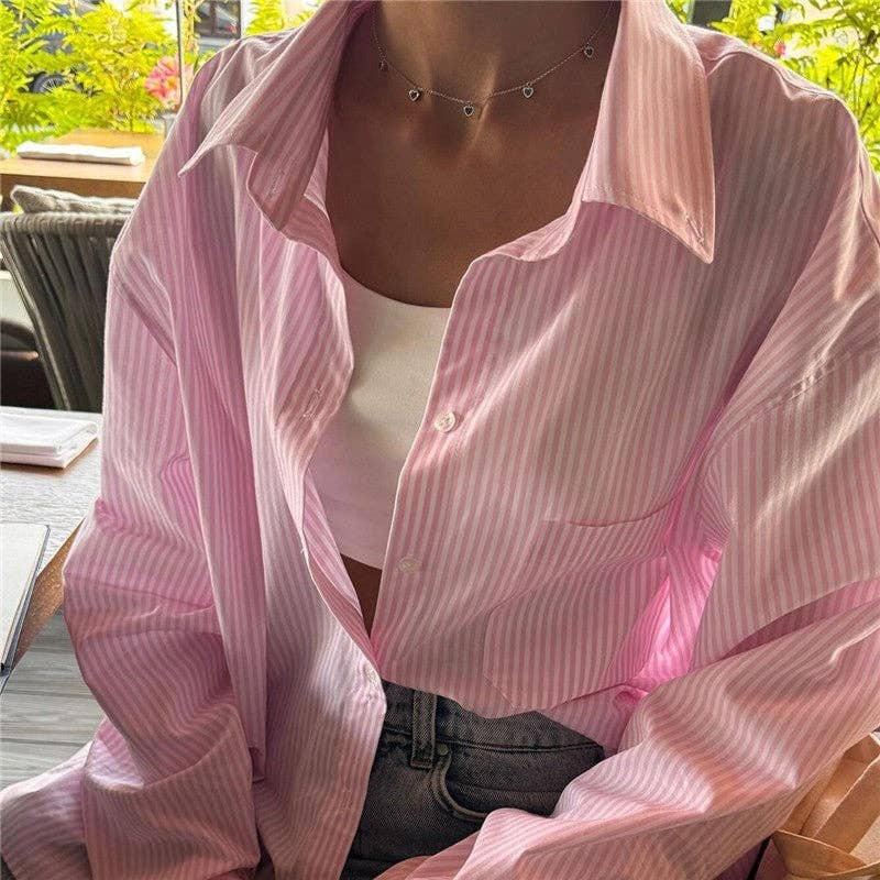 A person wearing an oversized pink and white vertically striped button-up shirt over a white crop top and dark gray jeans, photographed in a real indoor setting with visible furniture and greenery in the background.