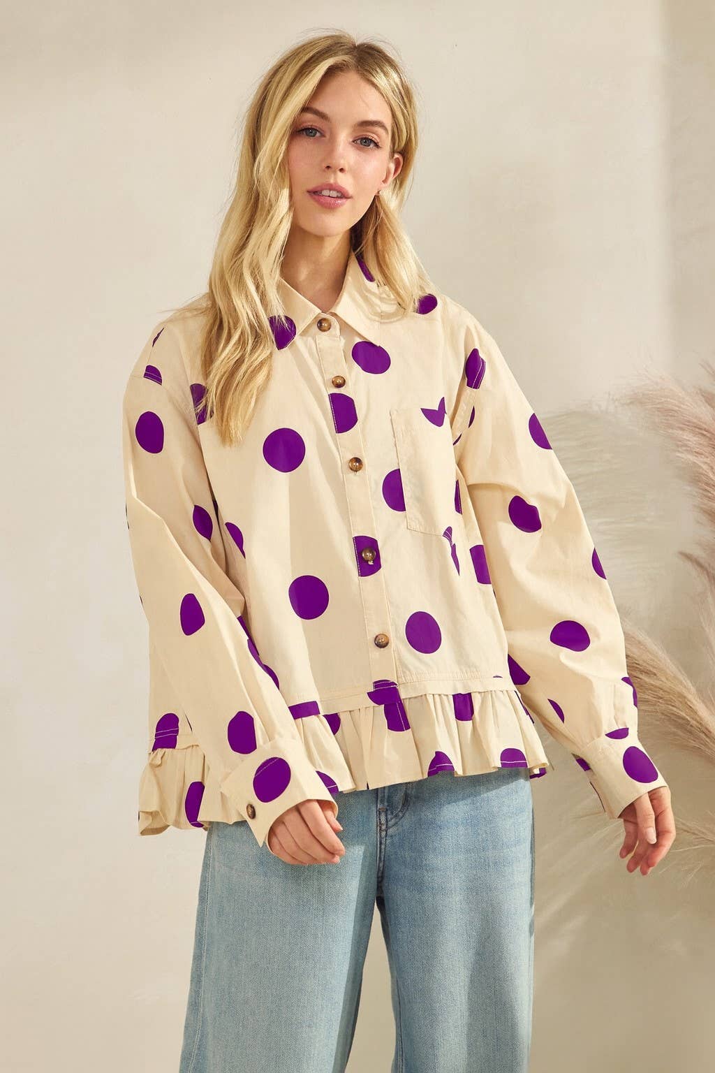 Woman wearing a beige blouse with purple polka dots and light blue jeans against a neutral background