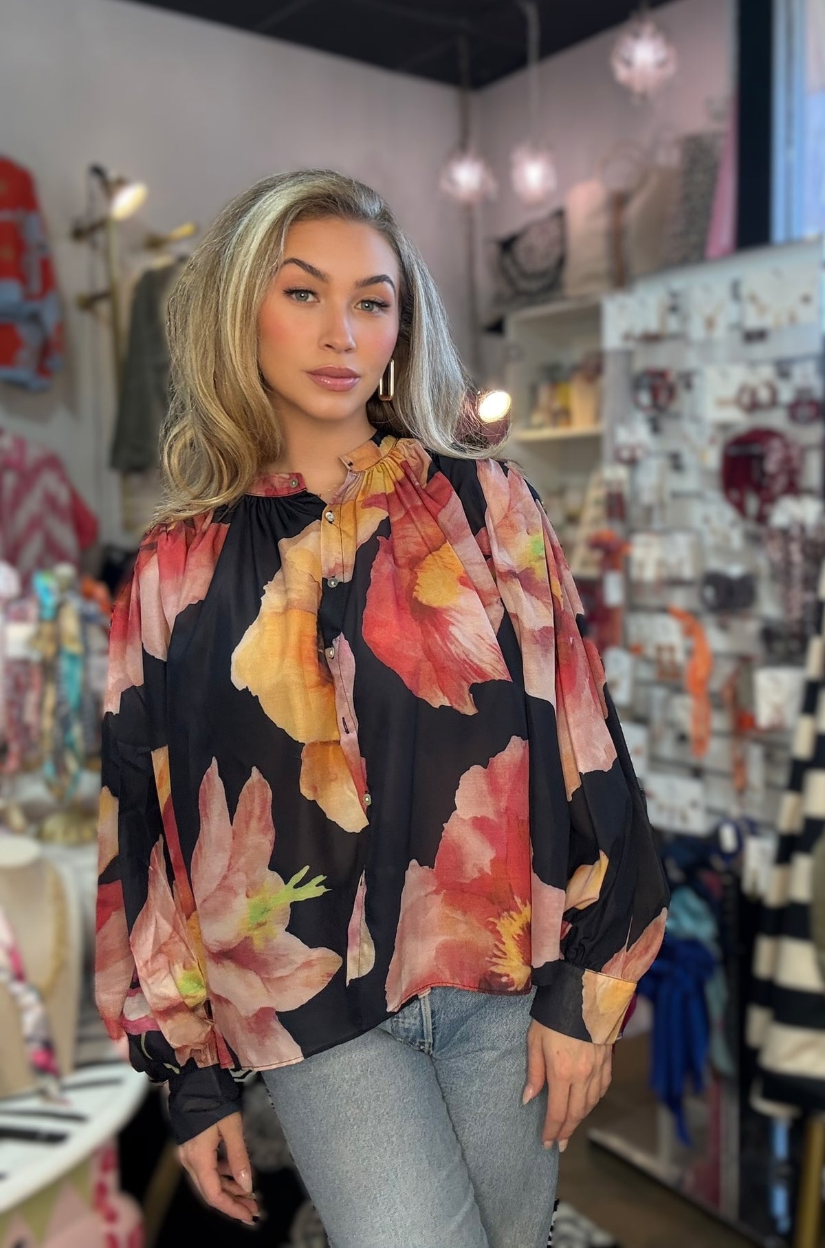 A woman with blonde hair wearing a black long-sleeve blouse with a vibrant pink, orange, and yellow floral print, paired with light blue jeans, standing inside a boutique.