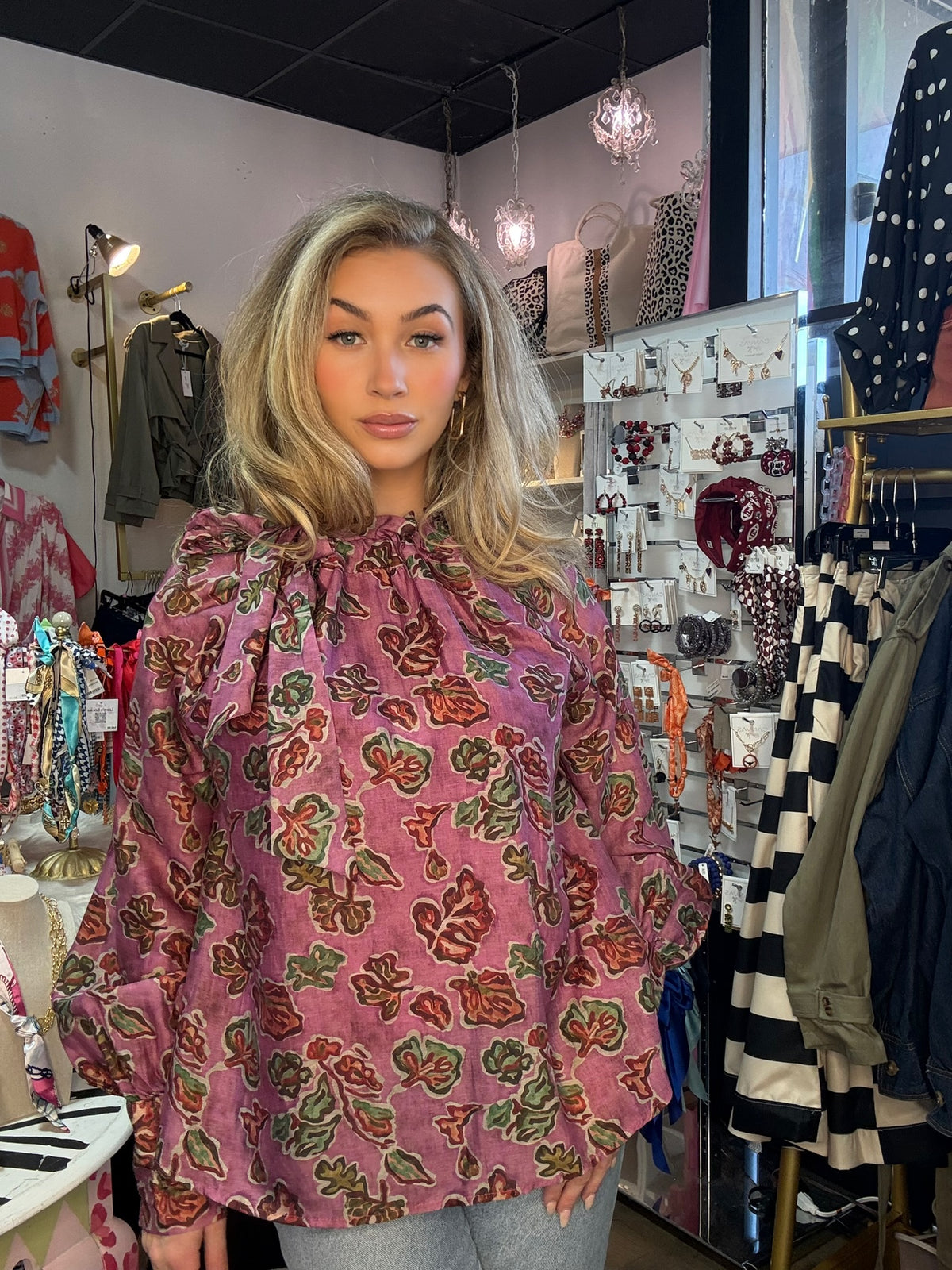 A woman with blonde hair wearing a pink long-sleeve blouse with a green and brown floral pattern, standing inside a clothing boutique.