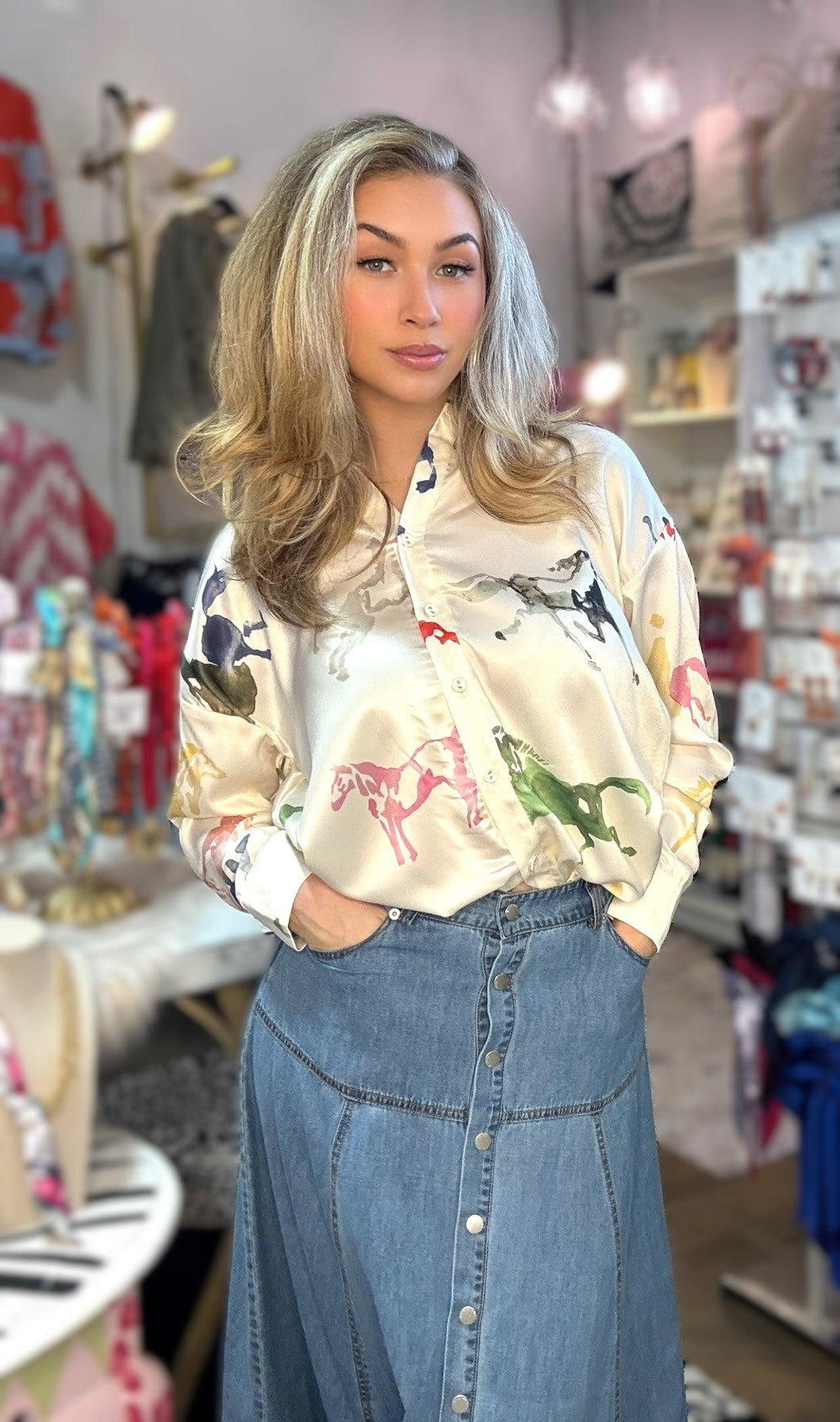 A woman with blonde-highlighted hair stands in a boutique wearing a cream satin button-up blouse with a colorful horse pattern and a blue denim button-front skirt.