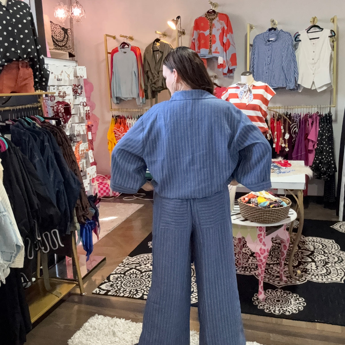 A person stands with their back to the camera inside a clothing boutique, wearing a matching blue ribbed long-sleeve top and wide-leg pants set. The shop is filled with racks of various garments, mannequins, and patterned rugs.