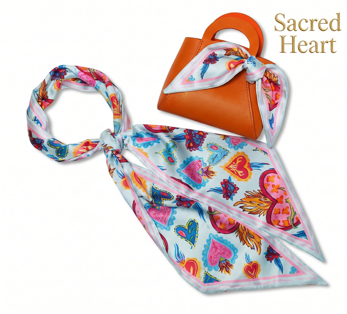 A colorful silk scarf with a heart and floral pattern, styled as a knot and draped next to a small orange handbag with a matching scarf, on a white background with 'Sacred Heart' text in the top right.