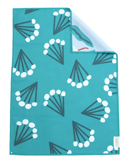 A square teal fabric item, likely a towel or blanket, featuring a repeating pattern of dark gray and white fan-shaped floral motifs. The top-right corner is folded over to show the reverse side, which has a light blue background with a small red graphic and a white brand tag.